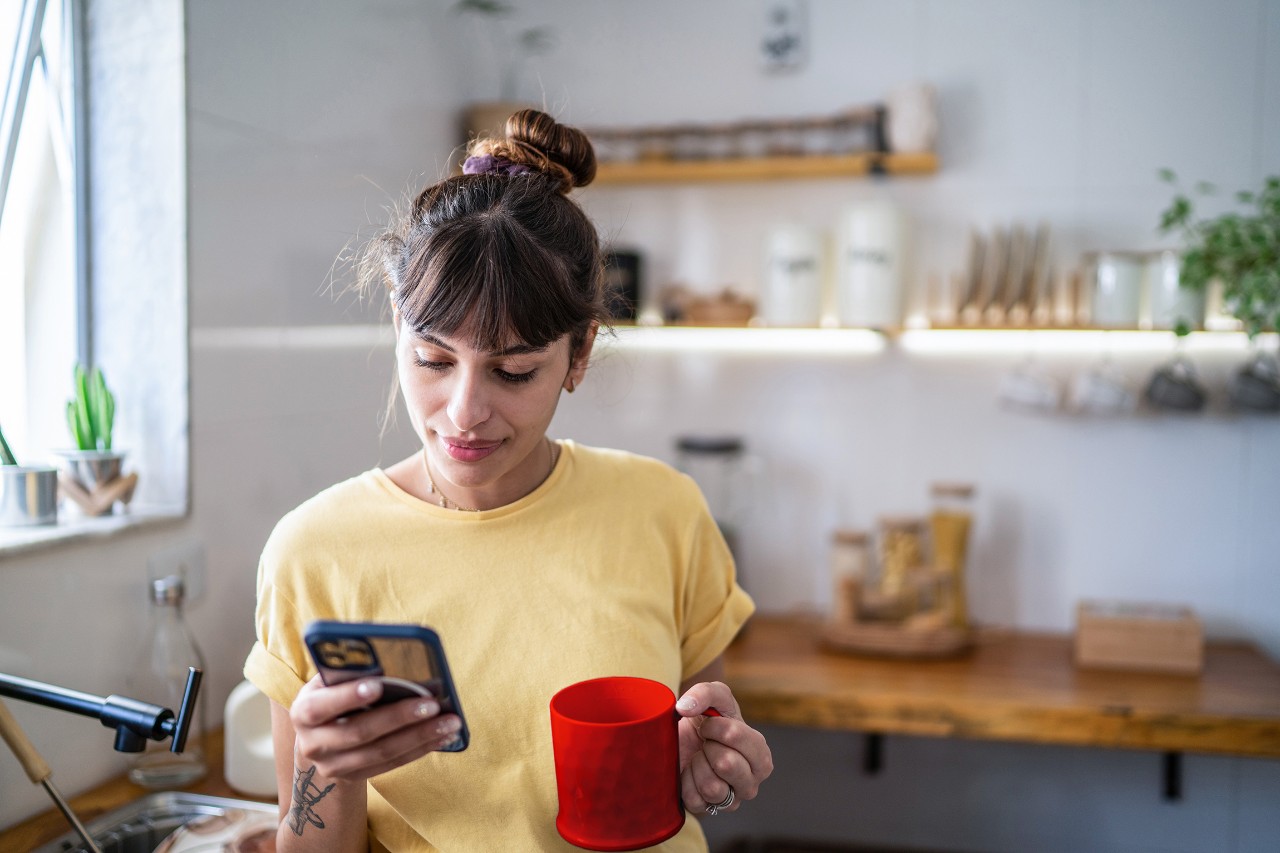 Femme dans la cuisine avec un smartphone et une tasse à la main